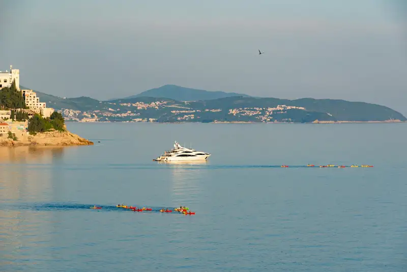 Scenic view of a luxury yacht and kayakers by the Dubrovnik coastline, perfect for summer travel and tourism.