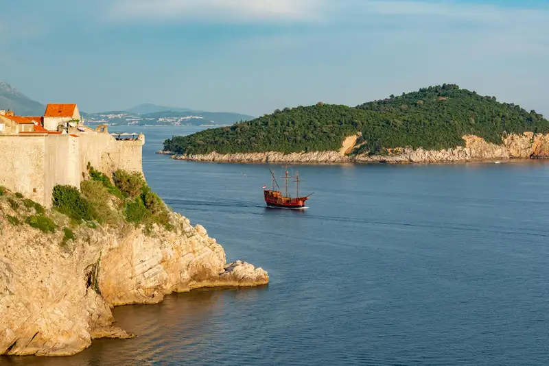 A picturesque view of Dubrovnik's historic coastline with an old ship sailing near a rocky cliff.
