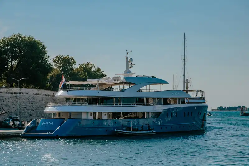 A luxurious yacht docked in a sunny harbor with trees and blue sky, capturing a serene summer day.