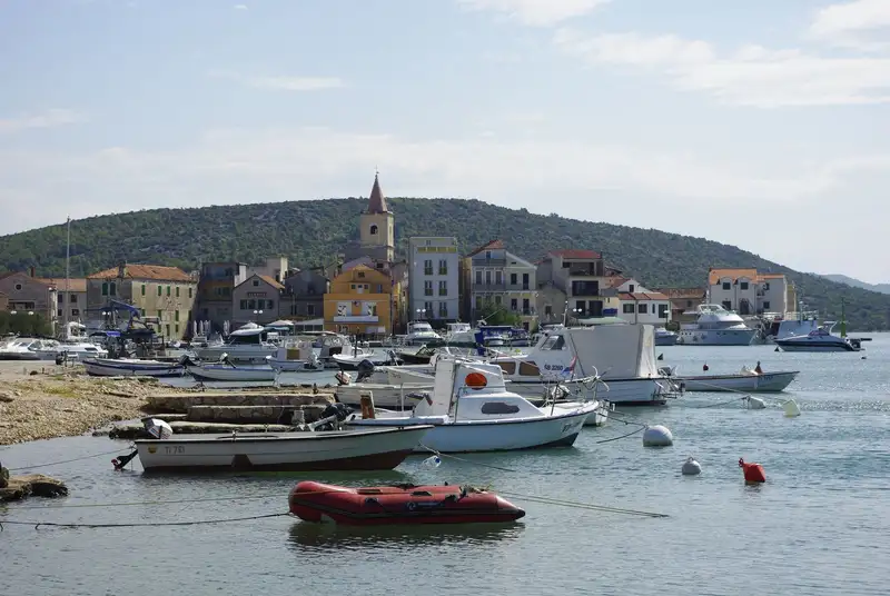 Boats docked at Pirovac harbor with hills and colorful buildings, Croatia.
